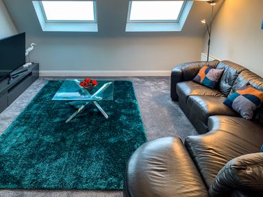 A living room with a glass coffee table on a teal rug and a black leather sofa under skylight windows at The Gables Penthouse in Bournemouth