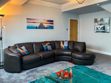 A living room with a curved leather sofa and colorful pillows a glass coffee table and wall art at The Gables Penthouse in Bournemouth