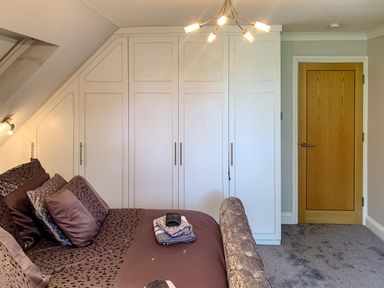 A bedroom with a bed, pillows, folded towels, built-in white wardrobes, a wooden door, and a ceiling light at The Gables Penthouse in Bournemouth