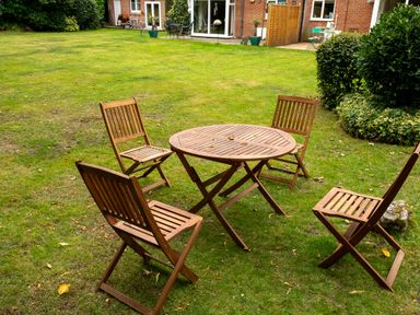 A wooden table with four wooden chairs set on a grassy lawn near a brick building with windows and a wooden fence