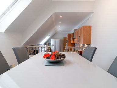 A dining area with a white table and gray chairs overlooking a kitchen with wooden cabinets at The Gables Penthouse in Bournemouth