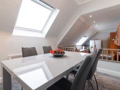 A dining area with a white table and four grey chairs next to a kitchen with wooden cabinets and a refrigerator at The Gables Penthouse in Bournemouth