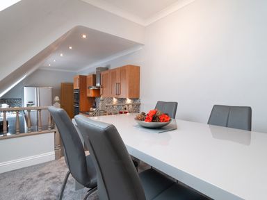 A dining area with a white table and gray chairs next to a kitchen with wooden cabinets at The Gables Penthouse in Bournemouth