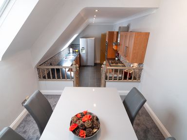 A dining table with four chairs and a bowl of pinecones and flowers overlooking a kitchen with wooden cabinets and a refrigerator at The Gables Penthouse in Bournemouth
