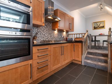 A kitchen with wooden cabinets stainless steel oven and hob with a tiled backsplash and a dining area up carpeted steps at The Gables Penthouse in Bournemouth
