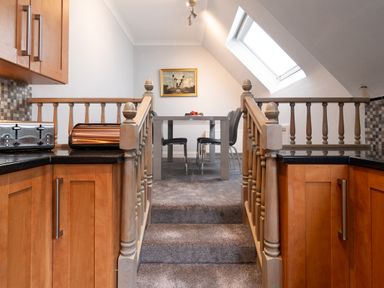 A kitchen area with wooden cabinets and black countertops leading up two carpeted steps to a dining area with a gray table and chairs under a skylight at The Gables Penthouse in Bournemouth