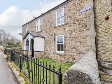 A stone house with white framed windows and a small porch behind a black metal fence at Tyne View Cottage in Bellingham