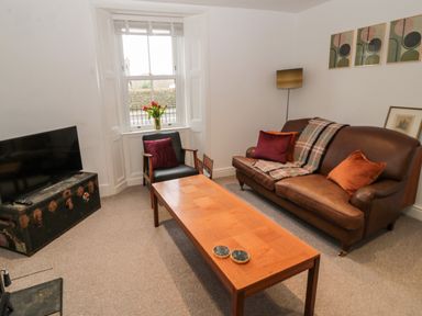 A living room with a brown leather sofa, wooden coffee table, armchair, TV on a trunk, and window with flowers at Tyne View Cottage in Bellingham
