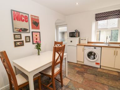 A kitchen with a white table and wooden chairs a washing machine a microwave and framed pictures on the wall at Tyne View Cottage in Bellingham