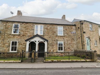 Stone cottage with multiple windows and black front doors behind black metal fence at Tyne View Cottage in Bellingham