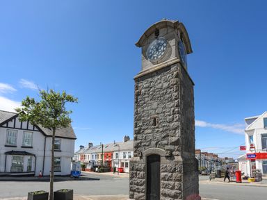 A stone clock tower on a street corner with houses and shops at Wingelock in Rhosneigr