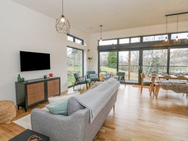 A living room with a gray sofa blue armchairs a wall-mounted TV and a dining table by large windows at Coorie In in Aberfeldy