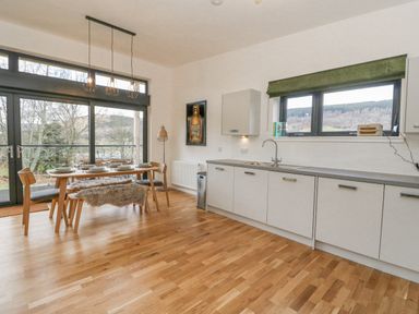A kitchen and dining area with wooden floor a dining table with chairs and a bench next to glass doors and cabinets with a sink under a window at Coorie In Aberfeldy