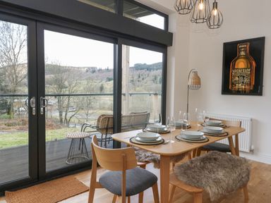 A dining area with a wooden table set with plates and glasses and large windows showing an outdoor seating area and countryside view at Coorie In in Aberfeldy