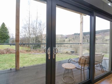 A patio with wicker chairs and a small round table outside glass doors overlooking trees and hills at Coorie In in Aberfeldy