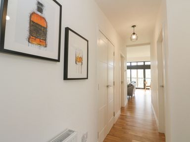 A hallway with wooden floor two framed pictures on the left wall and doors on both sides leading to a living room visible at the end at Coorie In Aberfeldy