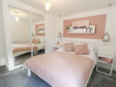 A bedroom with a white bed pink blanket two bedside tables and mirrored wardrobe at Coorie In Aberfeldy