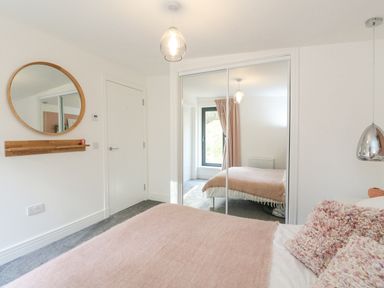 A bedroom with a bed covered with a pink blanket and pillows a round wall mirror and sliding mirrored closet doors at Coorie In Aberfeldy