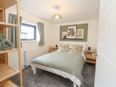 A bedroom with a bed covered with green bedding two nightstands and a shelving unit with towels at Coorie In Aberfeldy
