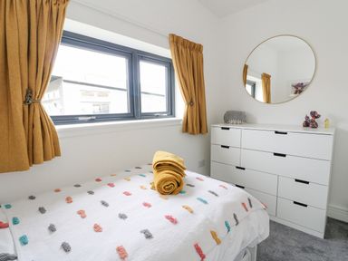 A bedroom with a bed covered with a blanket with colorful dots a white dresser with a round mirror and gold curtains at Coorie In Aberfeldy