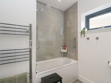 A bathroom with a bathtub shower combination a black stool a towel rack and a window at Coorie In Aberfeldy