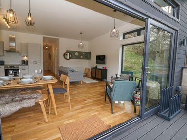 An open living and dining area with wooden flooring a dining table set with plates and glasses a sofa a small cabinet with a wall-mounted TV chairs and large glass sliding doors at Coorie In Aberfeldy