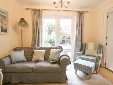 A living room with a sofa, armchair with footstool, floor lamp, and glass doors opening to a garden at The Old Lace Shop in Beer