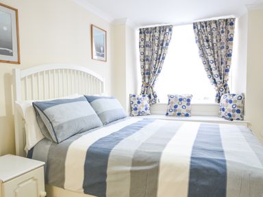 A bedroom with a bed with striped bedding and pillows next to a window with patterned curtains and cushions at The Old Lace Shop in Beer