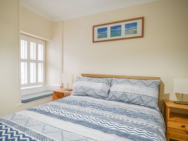 A bedroom with a bed featuring blue and white striped bedding two wooden bedside tables with lamps and a window with shutters at The Old Lace Shop in Beer