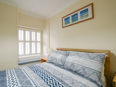 A bedroom with a bed with patterned bedding a wooden bedside table with a lamp and a window with white shutters at The Old Lace Shop in Beer