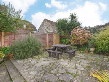 An outdoor patio area with stone paving a wooden picnic table and benches surrounded by plants and wooden fencing at The Old Lace Shop in Beer