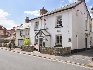 A street view of white buildings including Barrel Beer pub with a flag and two people walking on the pavement at The Old Lace Shop in Beer