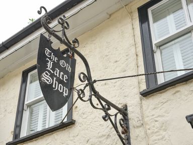 A black hanging sign with white text saying The Old Lace Shop on a beige building with two windows at The Old Lace Shop in Beer