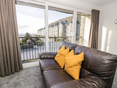 A living room with a brown leather sofa and yellow cushions by large windows with a view of buildings and a street at Borthwen in Barmouth