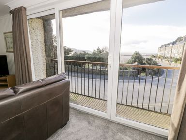 A living room with a brown leather sofa and glass doors leading to a balcony with a view of a street and buildings at Borthwen in Barmouth