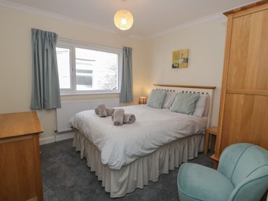 A bedroom with a double bed with rolled towels on it a wooden wardrobe a wooden chest of drawers a small green chair and curtains at Borthwen in Barmouth