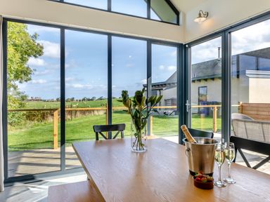 A dining area with a wooden table chairs a glass vase with flowers a champagne bucket two glasses and strawberries by large windows overlooking a green field at Shearwater Lodge in Bamburgh
