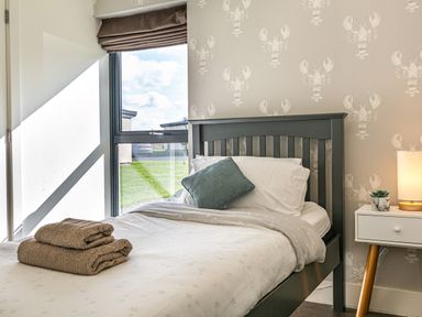 A bedroom with a single bed featuring white bedding and a green pillow next to a window and a nightstand with a lamp and plant at Shearwater Lodge in Bamburgh
