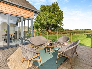 A wooden deck with outdoor seating and a small table with coffee press and mugs surrounded by glass doors and green fields at Shearwater Lodge in Bamburgh