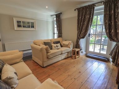 A living room with two beige sofas a small wooden stool and glass doors with curtains at Brook Cottage in Windermere