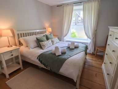 A bedroom with a bed with pillows and towels bedside tables lamps a window with curtains a dresser and a chair at Brook Cottage in Windermere