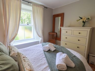 A bedroom with a bed towels on a bed a window with curtains a chair a mirror and a chest of drawers with flowers at Brook Cottage in Windermere