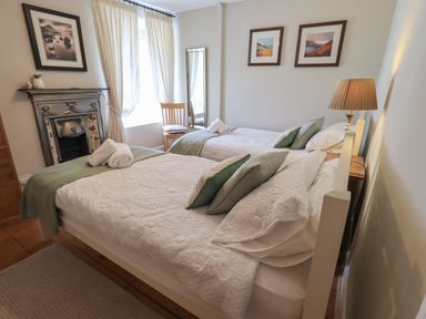A bedroom with two single beds with white bedspreads and green pillows a wooden chair near window with white curtains a decorative fireplace and framed pictures on the wall at Brook Cottage in Windermere