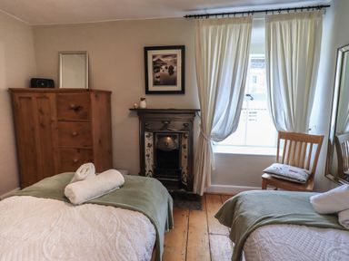 A bedroom with two single beds with white covers and green throws a wooden wardrobe a fireplace a chair with a cushion and a window with curtains at Brook Cottage in Windermere