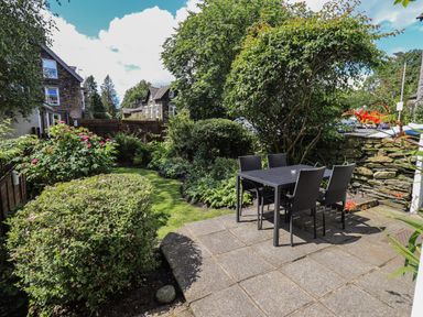 A garden with a patio area furnished with a table and four chairs surrounded by bushes and trees at Brook Cottage in Windermere