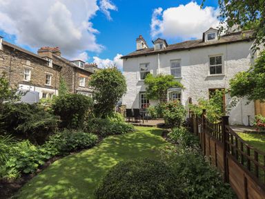 A garden with plants and a patio area with outdoor furniture behind a white house at Brook Cottage in Windermere