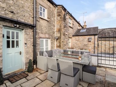 An outdoor patio area with wicker furniture and glass table in front of a stone house at Market Square Maisonette in Kirkby Lonsdale