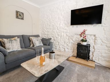 A living room with a gray sofa a marble coffee table a wall-mounted television and a small stove with flowers at Market Square Maisonette in Kirkby Lonsdale