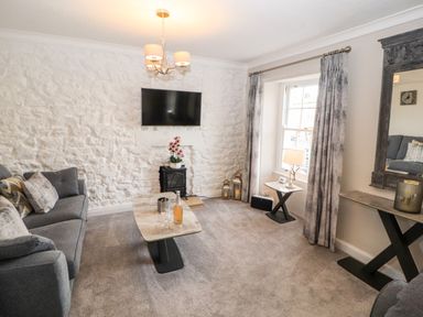 A living room with a grey sofa a marble coffee table a television mounted on a white stone wall and a window with grey curtains at Market Square Maisonette in Kirkby Lonsdale