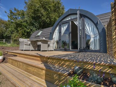 An outdoor wooden deck with wicker chairs and table outside a pod-shaped building with large glass doors at Maple in Wootton Fitzpaine near Charmouth